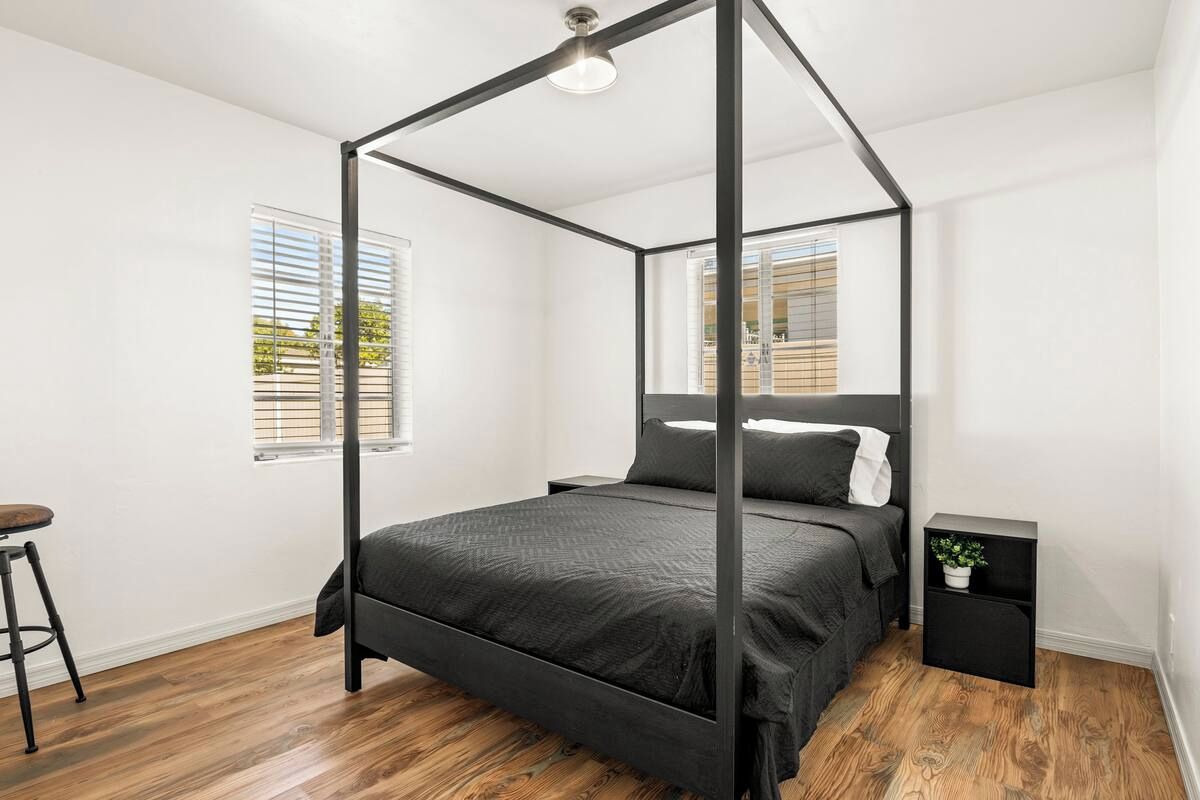 A modern bedroom with a four-poster bed, a small nightstand, and a window with blinds.