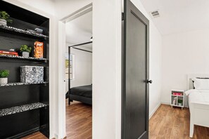 A bedroom with a bed, a white bedside table, a shelf with books and decorative items, and a door leading to another room. - 2 Bedroom in WPB - Spacious Backyard - Prime Location (Unknown)