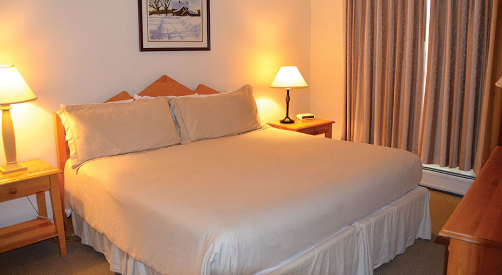 A neatly made bed with a headboard, two bedside lamps, a framed picture on the wall, and a window with curtains.