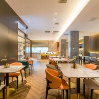 Un restaurante moderno con varias mesas y sillas, una zona de cocina al fondo y un suelo con estampado de espiga.