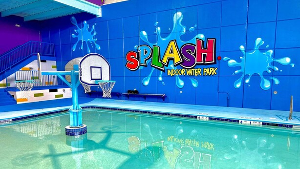 Un parc aquàtic interior amb un cèrcol de bàsquet, una piscina i una paret blava amb la inscripció 'SPLASH INDOOR WATER PARK'.