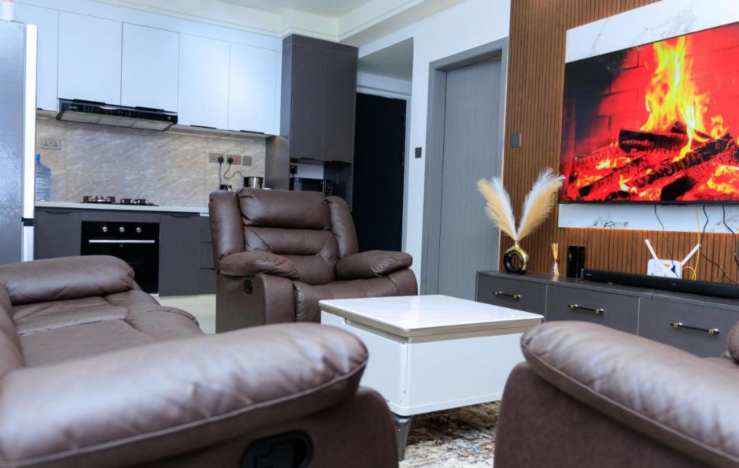 A modern living room with leather sofas, a flat-screen TV, and a kitchen in the background.