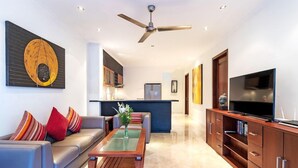 A modern living room with a sofa, coffee table, and TV. There is a kitchen in the background with a refrigerator and cabinets.