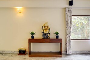 Una estàtua sobre una taula de consola de fusta, amb dues plantes en testos a la vora, una làmpada penjada a la paret i una finestra amb cortines.