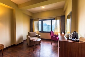 A hotel room with a wooden floor, a sofa, a chair, a small round table, a desk with a coffee maker, and a window with curtains. - Mount Himalayan Hotel & Spa Gangtok (Gangtok)