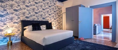 Un dormitorio con cama, mesita de noche, pared con estampado floral y una puerta que da a otra habitación.