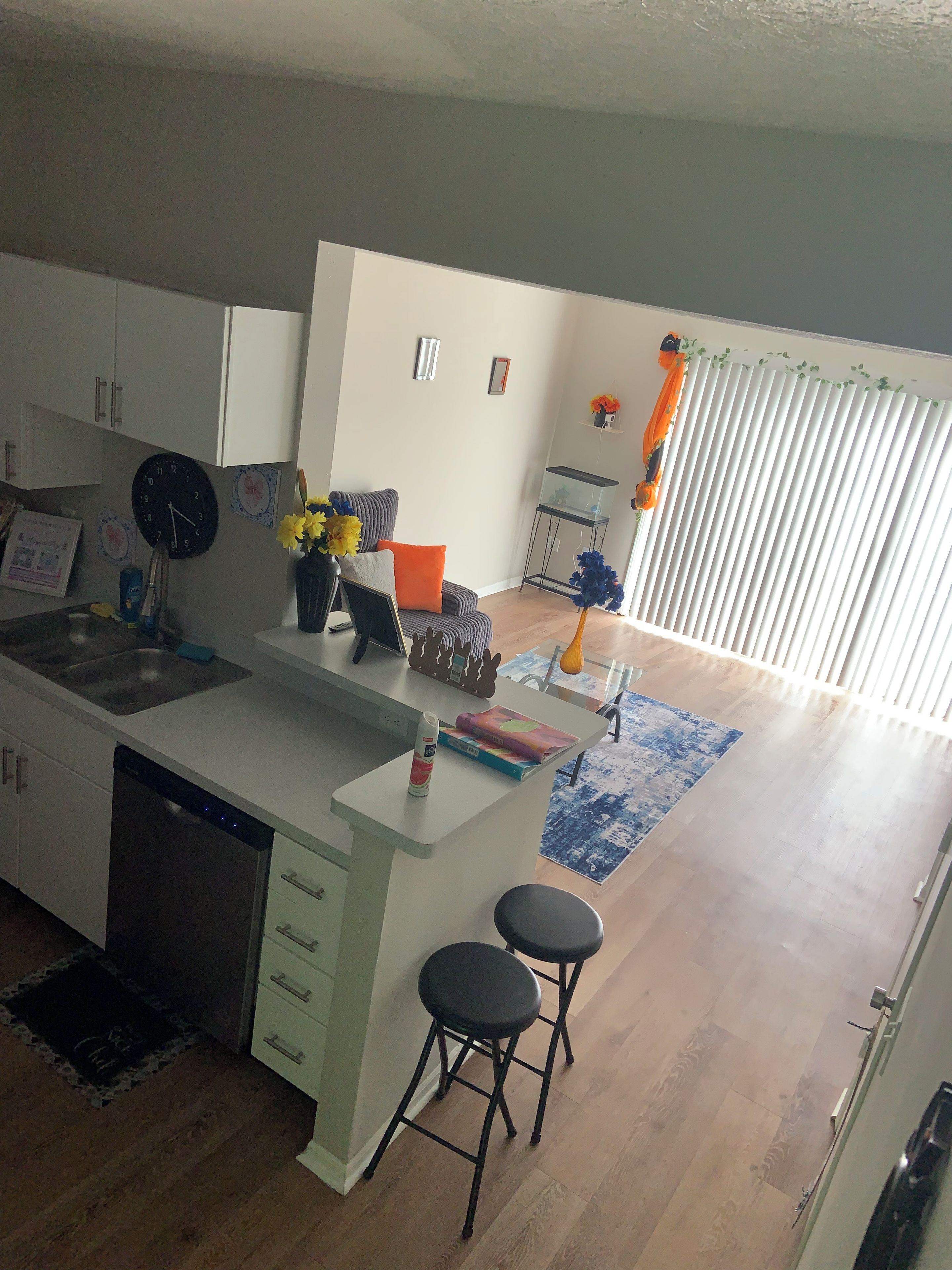 A modern kitchen with a dishwasher, a sink, and a built-in island with stools. There is a window with blinds and a decorative plant on the windowsill.
