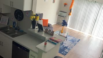 A modern kitchen with a dishwasher, a sink, and a built-in island with stools. There is a window with blinds and a decorative plant on the windowsill.