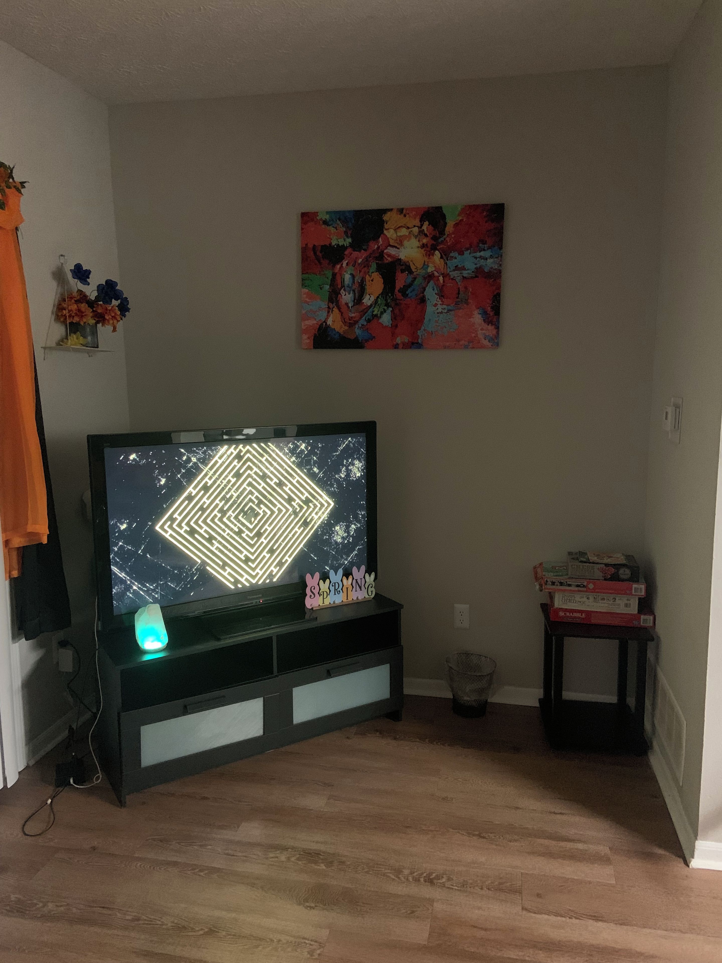 A living room with a flat-screen TV on a stand, a colorful abstract painting on the wall, and a small table with board games.