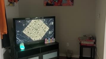 A living room with a flat-screen TV on a stand, a colorful abstract painting on the wall, and a small table with board games.