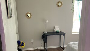 Un dormitorio con cama, escritorio con lámpara, mesita con toallas, espejo en la pared y ventana con cortinas.