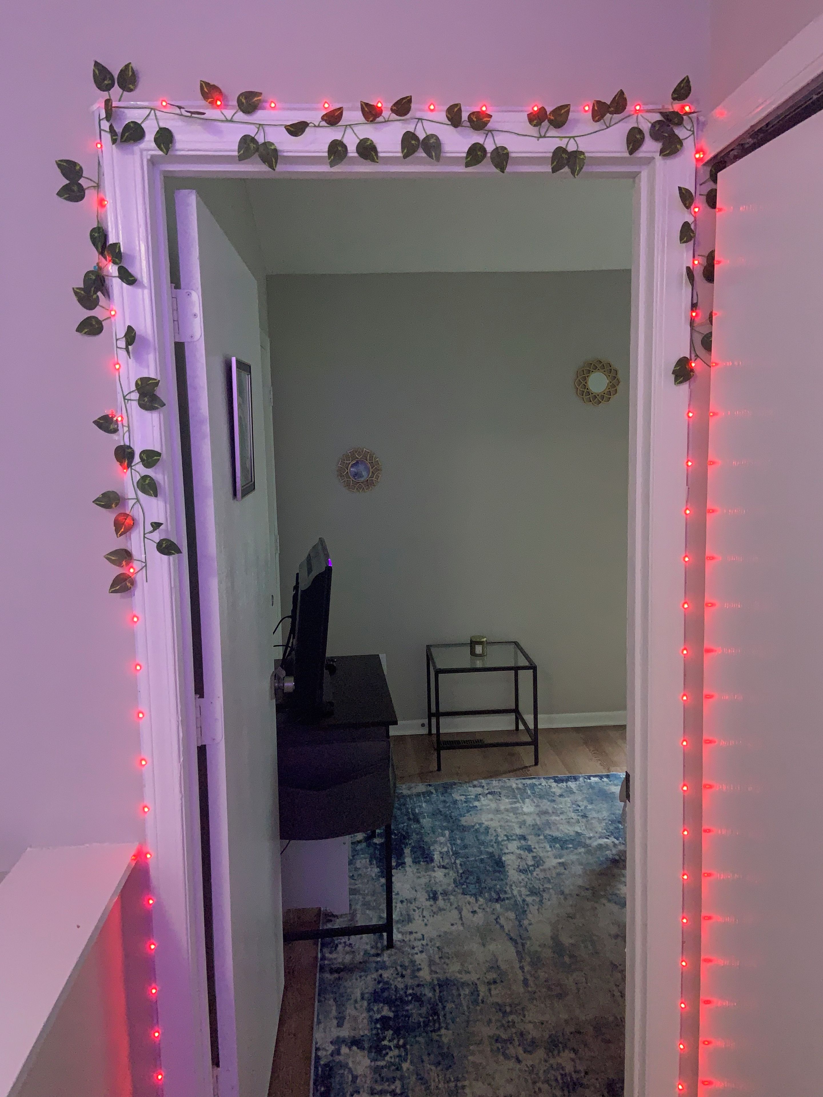 A doorway with a decorative garland and red LED lights, leading to a room with a desk, chair, and a small table with a decorative item.