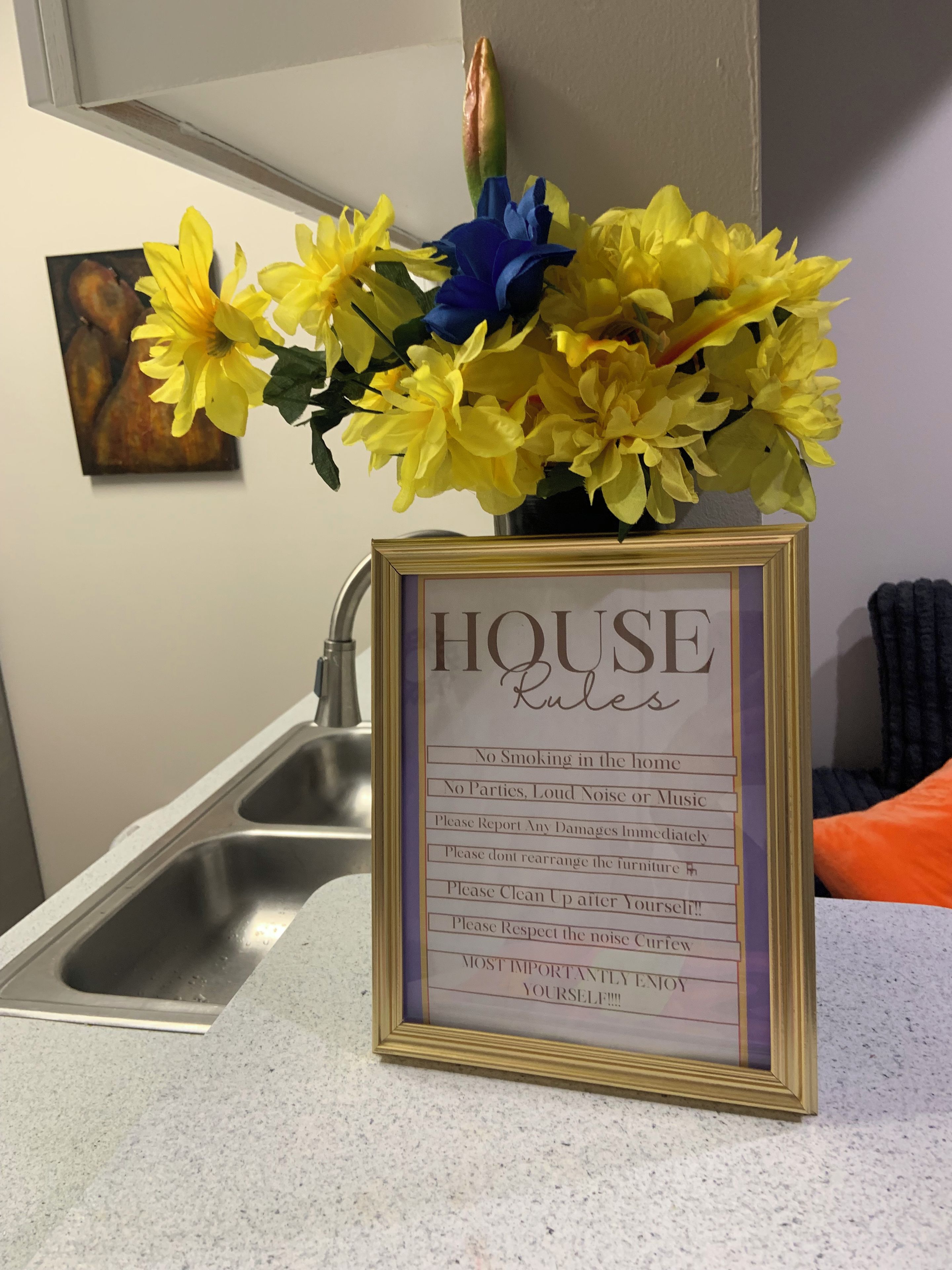 Ein gerahmtes Schild mit Hausordnung auf einer Küchenarbeitsplatte, einem Spülbecken und einer Vase mit gelben und blauen Blumen.