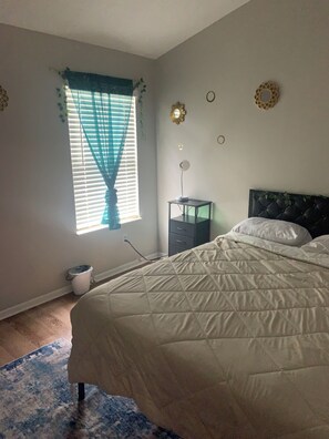 Un dormitorio con cama, mesita de noche, lámpara, ventana con cortinas y un adorno en la pared.
