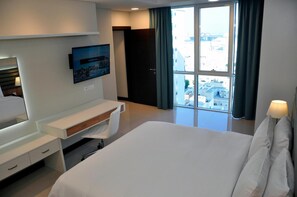 A hotel room with a bed, a desk, a TV mounted on the wall, and a large window with a view of buildings. - Gulf Executive Residence Juffair Gerj (Manama)