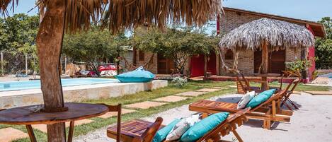 Un resort con sillones de madera y una sombrilla de paja, una piscina y un edificio de ladrillo al fondo.