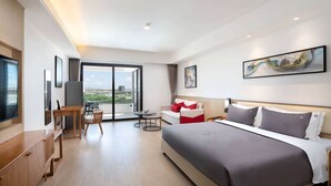 A modern hotel room with a bed, a desk, a chair, and a view through a large window. - Ramada Encore by Wyndham Jiusuo Downtown (Ledong)