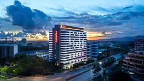 A multi-story hotel with a prominent sign, surrounded by other buildings and a clear sky. - Ramada Encore by Wyndham Jiusuo Downtown (Ledong)