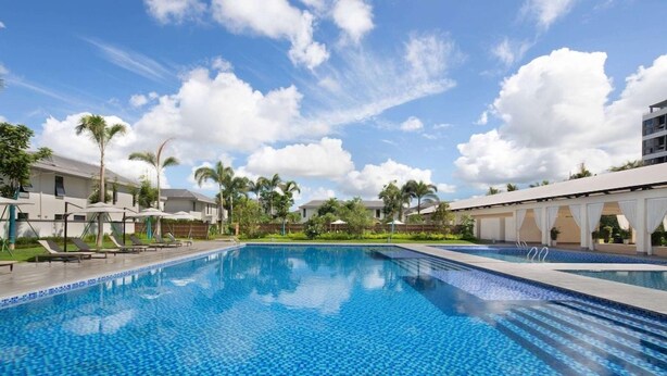 A swimming pool with lounge chairs and umbrellas, surrounded by buildings and palm trees. - Ramada Encore by Wyndham Jiusuo Downtown (Ledong)