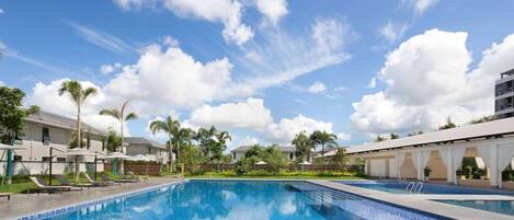 A swimming pool with lounge chairs and umbrellas, surrounded by buildings and palm trees.