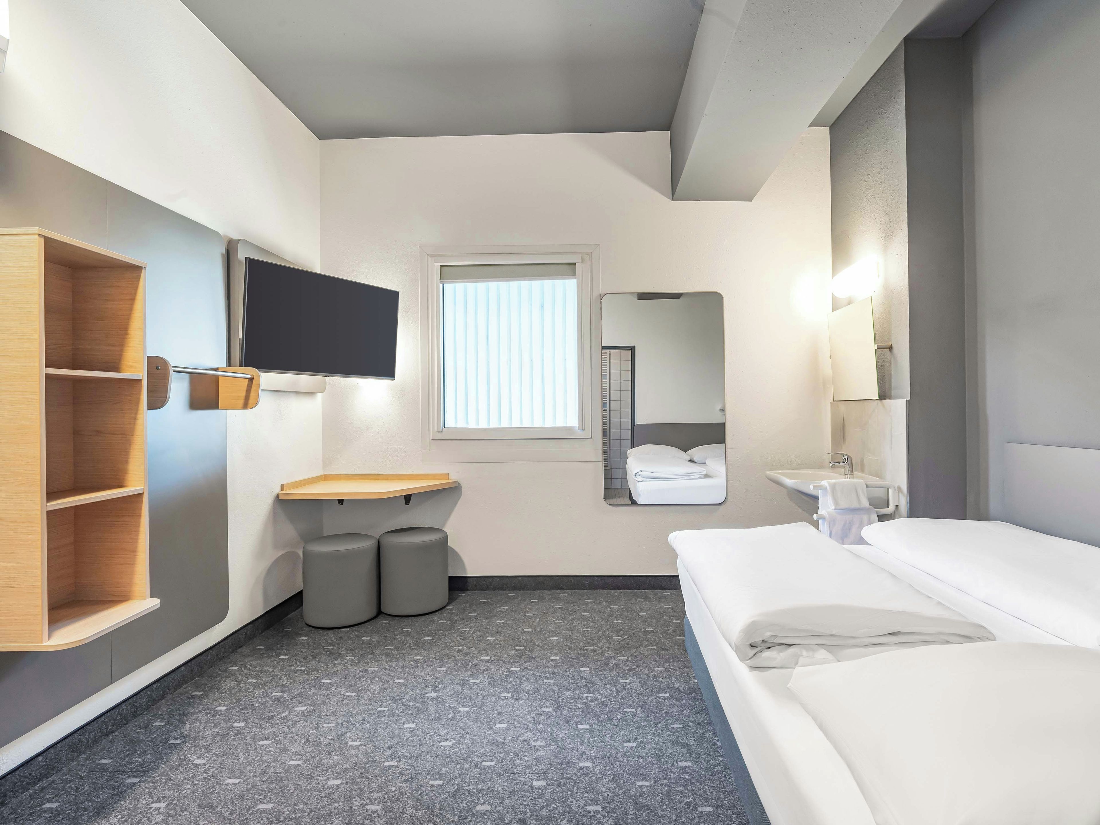 Ein Hotelzimmer mit Bett, Wandfernseher, kleiner Holzablage, Fenster mit Jalousien, Spiegel und Waschbecken.