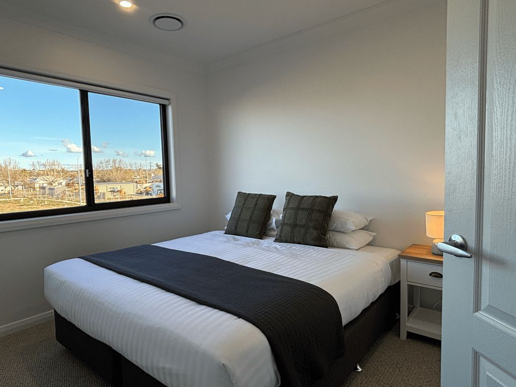 A bedroom with a large window, a bed with white bedding and a black throw, a bedside table with a lamp, and a door to the right.
