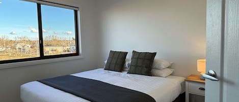 A bedroom with a large window, a bed with white bedding and a black throw, a bedside table with a lamp, and a door to the right.