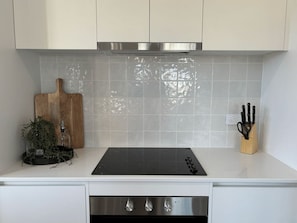 A kitchen with white cabinets, a stainless steel range hood, a black induction cooktop, a wooden cutting board, and a knife block. - Summons Forty - Sleeps 6 (Wagga Wagga)