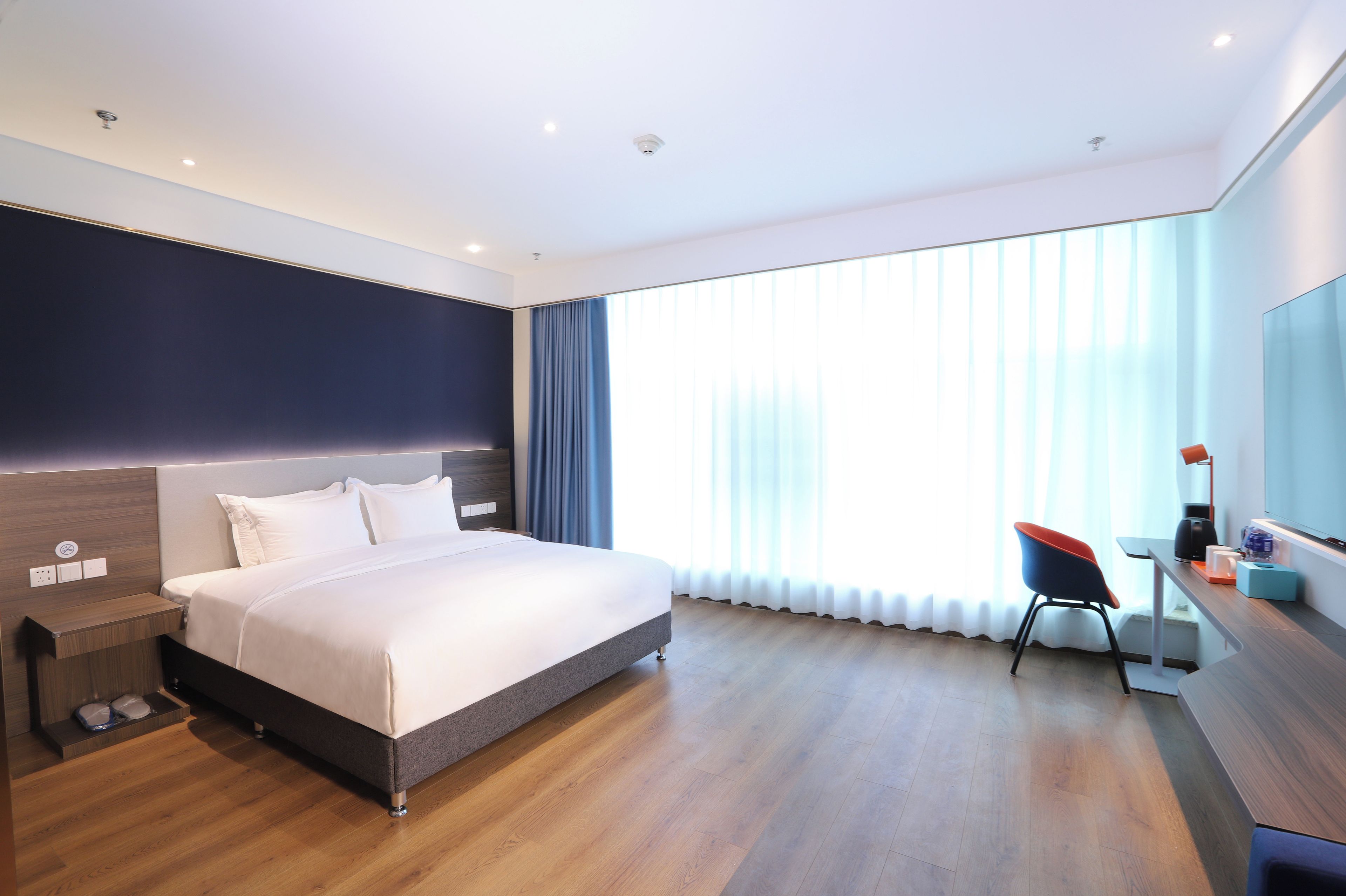 A modern hotel room with a large bed, a desk with a chair, and a large window with curtains.