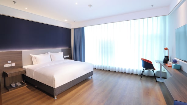 Quarto de hotel moderno com uma cama grande, uma escrivaninha com cadeira e uma janela ampla com cortinas.