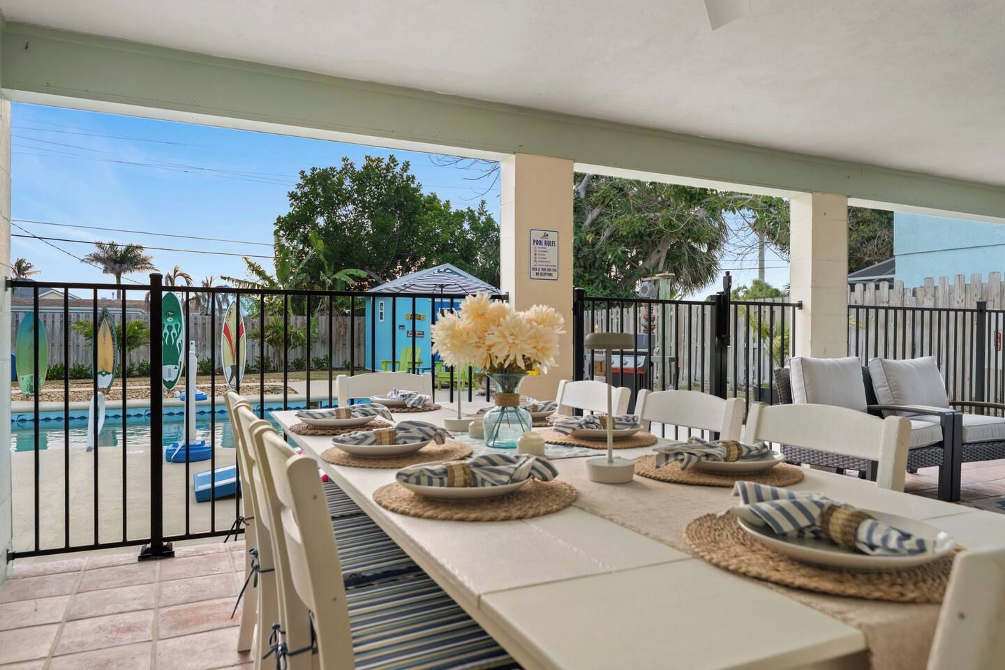 A covered outdoor dining area with a pool view, featuring a table set with plates and a vase of flowers.