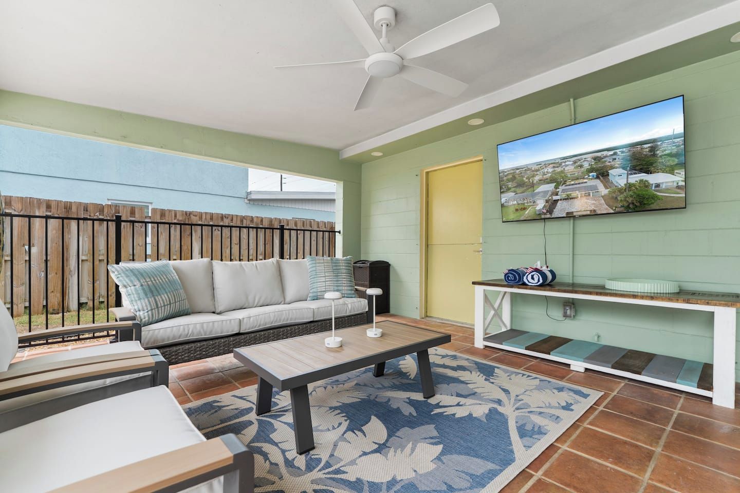 A room with a ceiling fan, a sofa with cushions, a coffee table, a TV mounted on the wall, and a view of the outside.