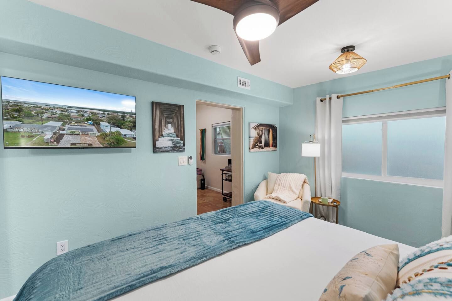 A bedroom with a bed, a window, a ceiling fan, and a wall-mounted TV displaying an aerial view of a residential area.
