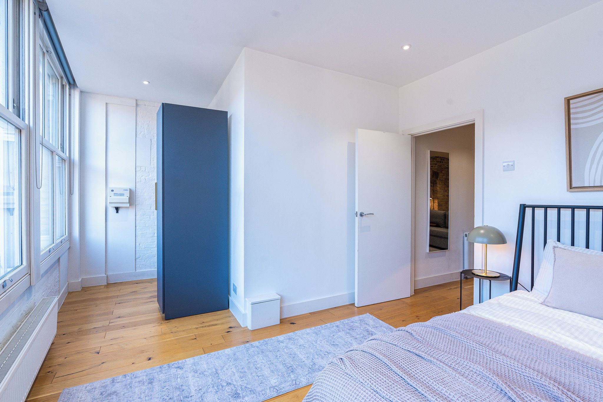 A modern bedroom with a large bed, a bedside table with a lamp, a window with blinds, and a door leading to another room.