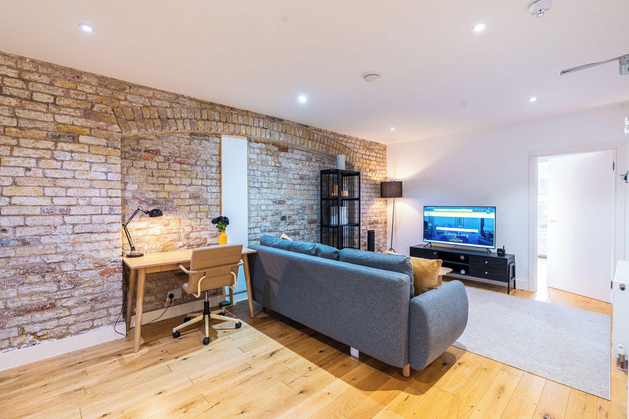 Een moderne woonkamer met een bakstenen muur, houten vloer, een grijze bank, een houten bureau met een stoel, een televisie en een zwarte boekenkast.