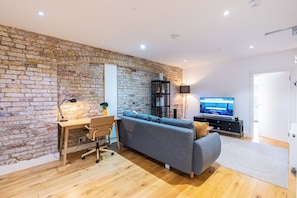 A modern living room with a brick wall, wooden flooring, a grey sofa, a wooden desk with a chair, a TV, and a black shelving unit.