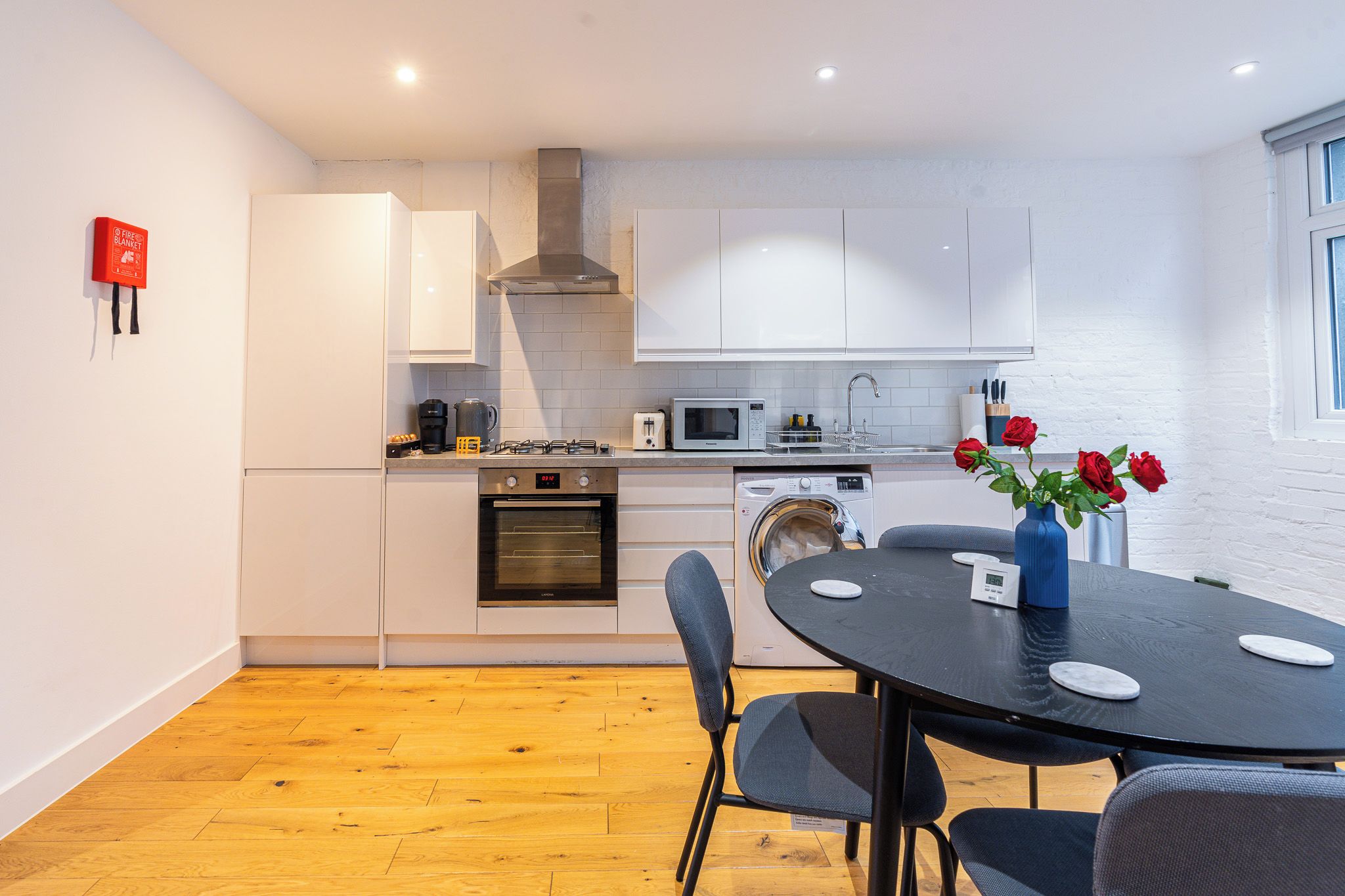 Een moderne keuken met witte kasten, een ingebouwde oven, magnetron en wasmachine. Er staat een eettafel met stoelen en een vaas met rode rozen.