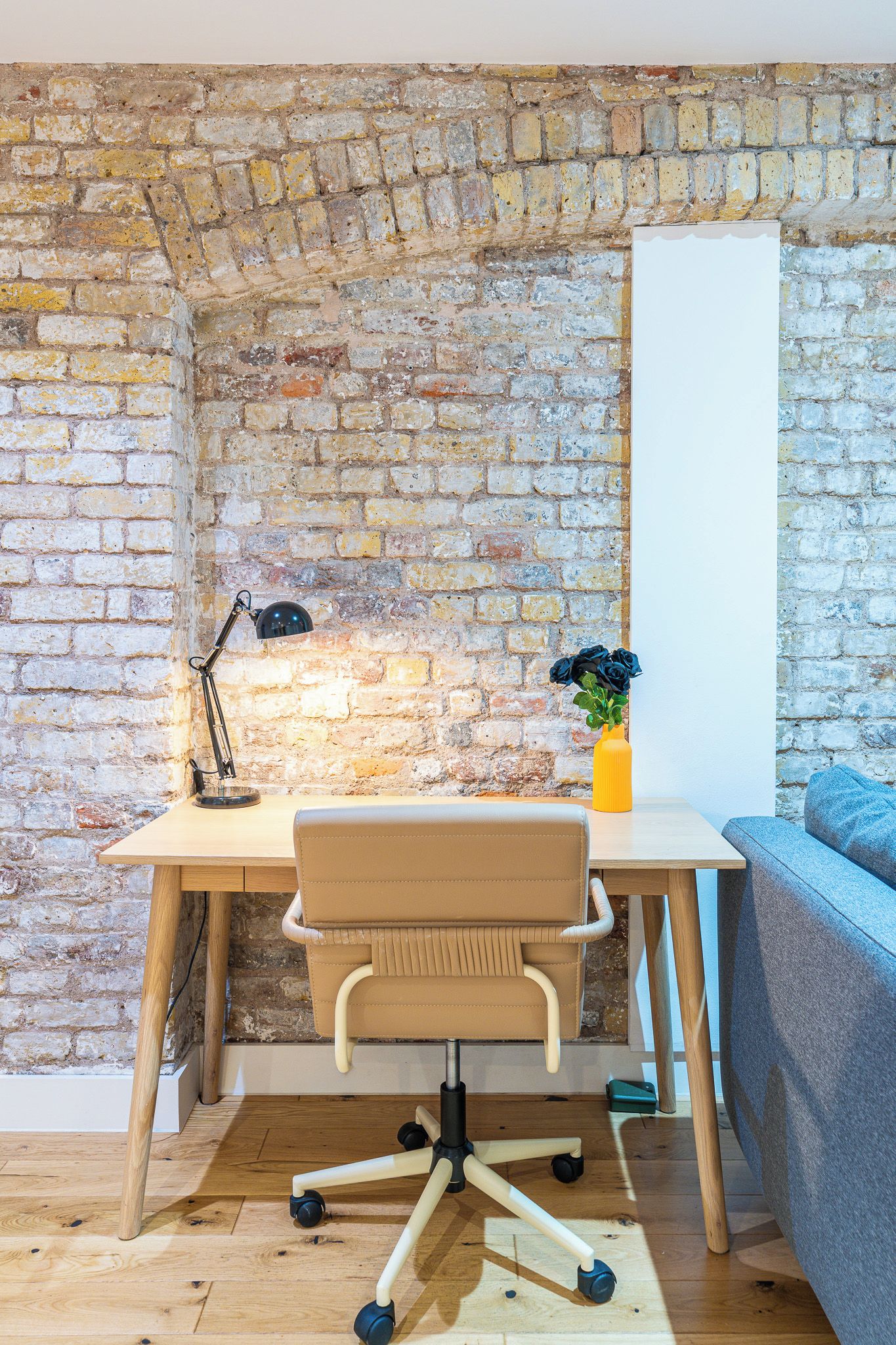 A corner office with a brick wall, a desk with a chair, a lamp, and a vase with flowers.