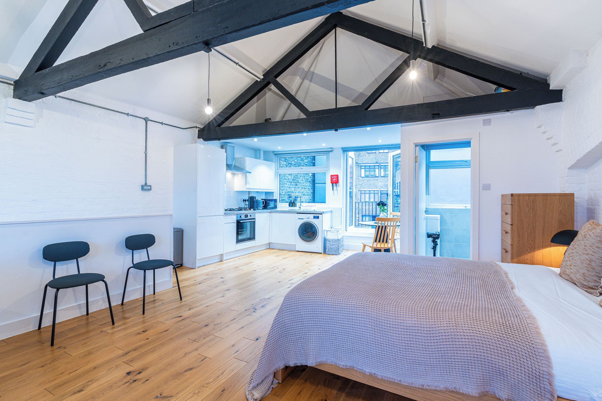 A modern loft bedroom with a large bed, a wooden ceiling, and a kitchen area visible through an open door.