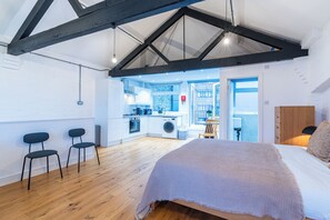 A modern loft bedroom with a large bed, a wooden ceiling, and a kitchen area visible through an open door.