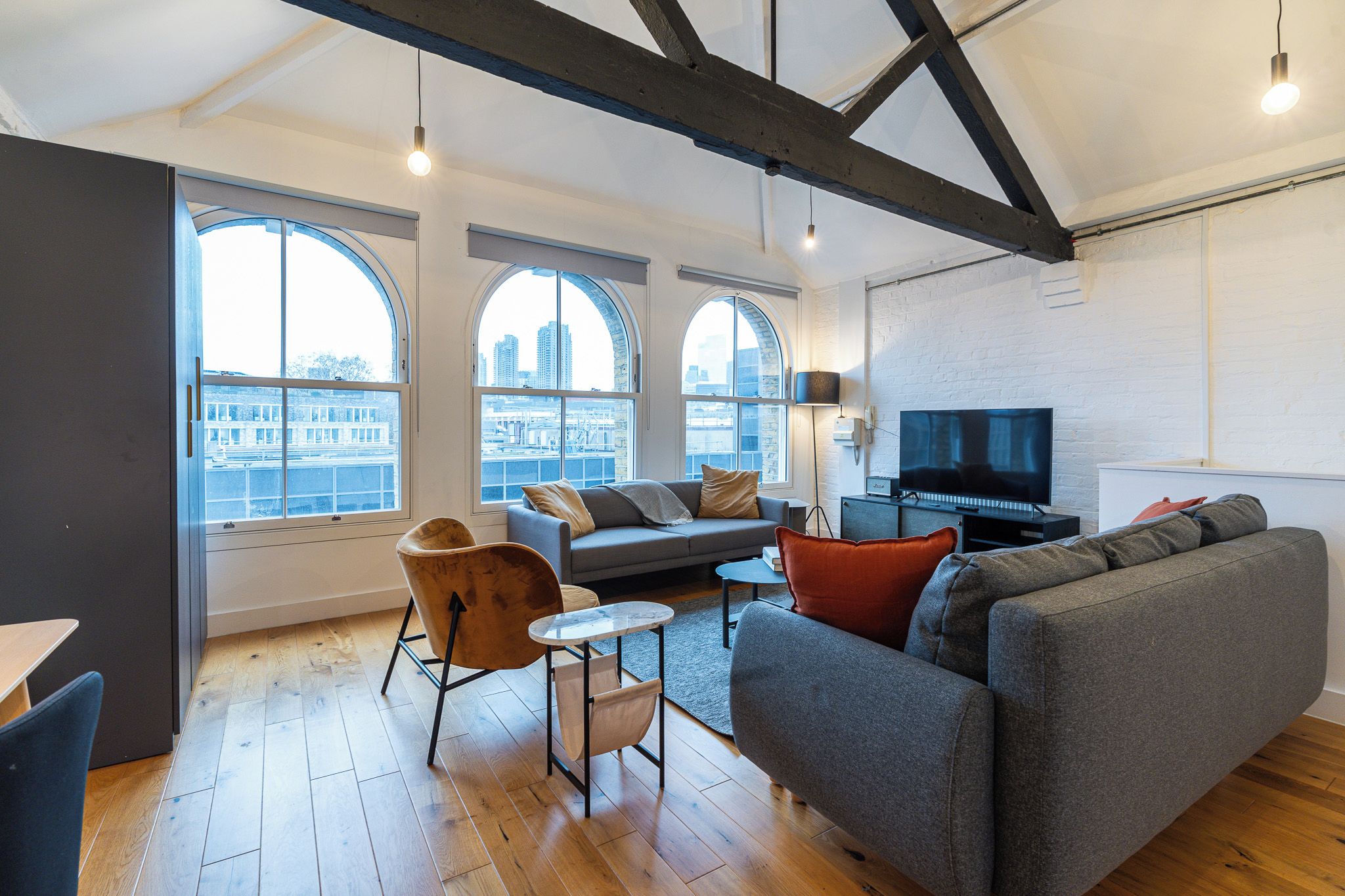 Un salon moderne comprenant un canapé, un fauteuil et un téléviseur, de grandes fenêtres offrant une vue sur la ville, et des poutres apparentes en bois.
