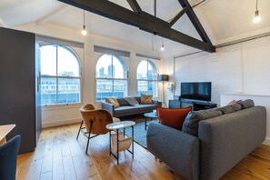 Un salon moderne comprenant un canapé, un fauteuil et un téléviseur, de grandes fenêtres offrant une vue sur la ville, et des poutres apparentes en bois.