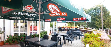 An outdoor seating area with tables and chairs, a covered section with a logo, and a building with large windows in the background.