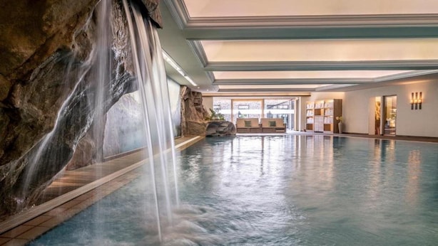 A large indoor swimming pool with a waterfall feature, a reception area with seating, and a view of the outdoors. - Hotel Franks (Oberstdorf)