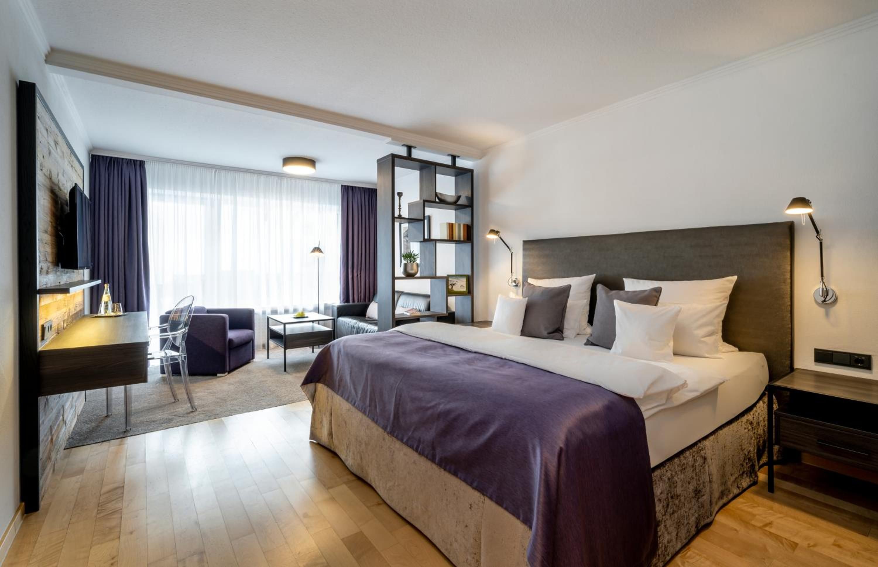 Ein modernes Hotelzimmer mit einem großen Bett, einem Schreibtisch, einem Sessel, einem Bücherregal und einem Fernseher.