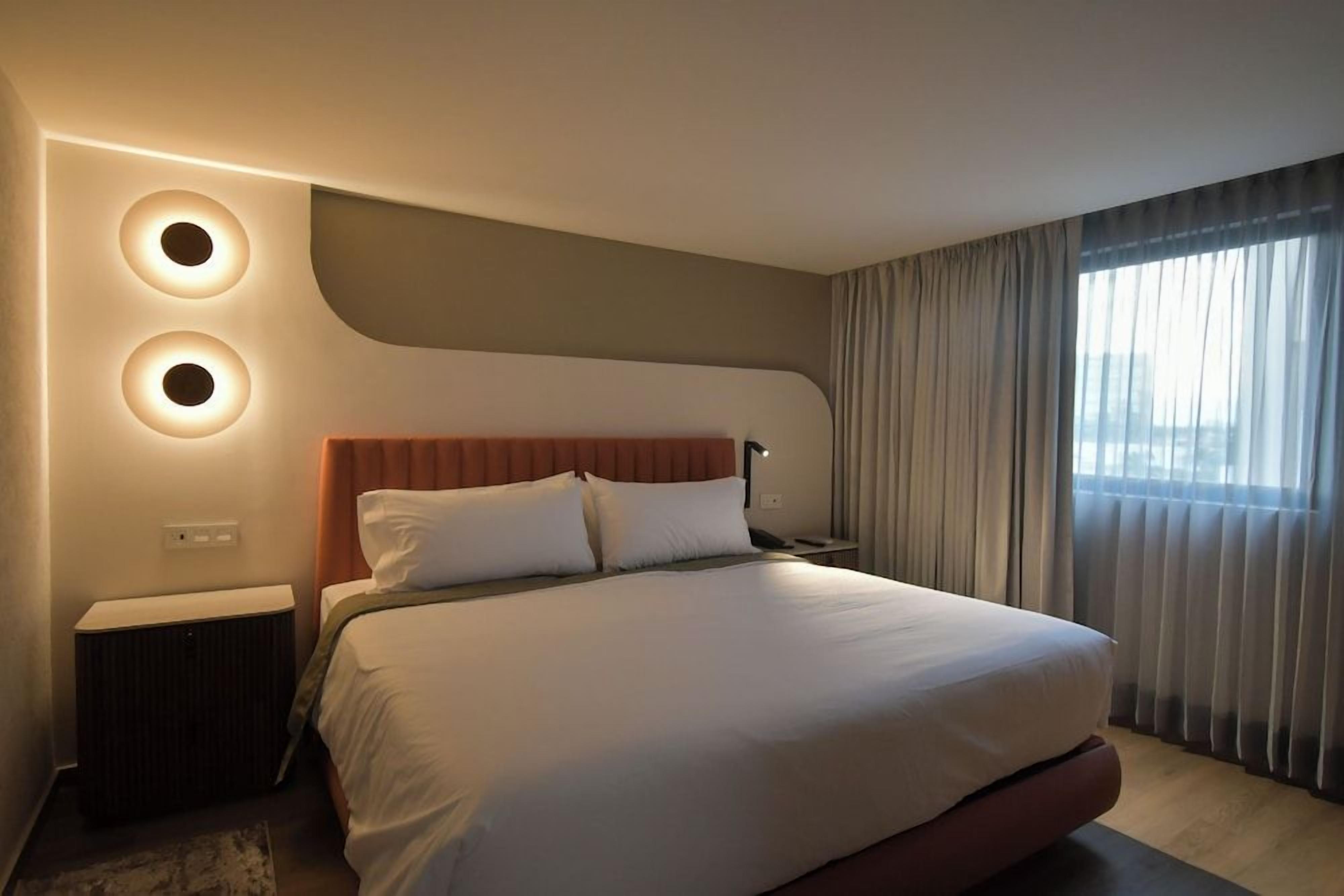 A modern hotel room with a large bed, a nightstand, a wall-mounted lamp, and a window with curtains.