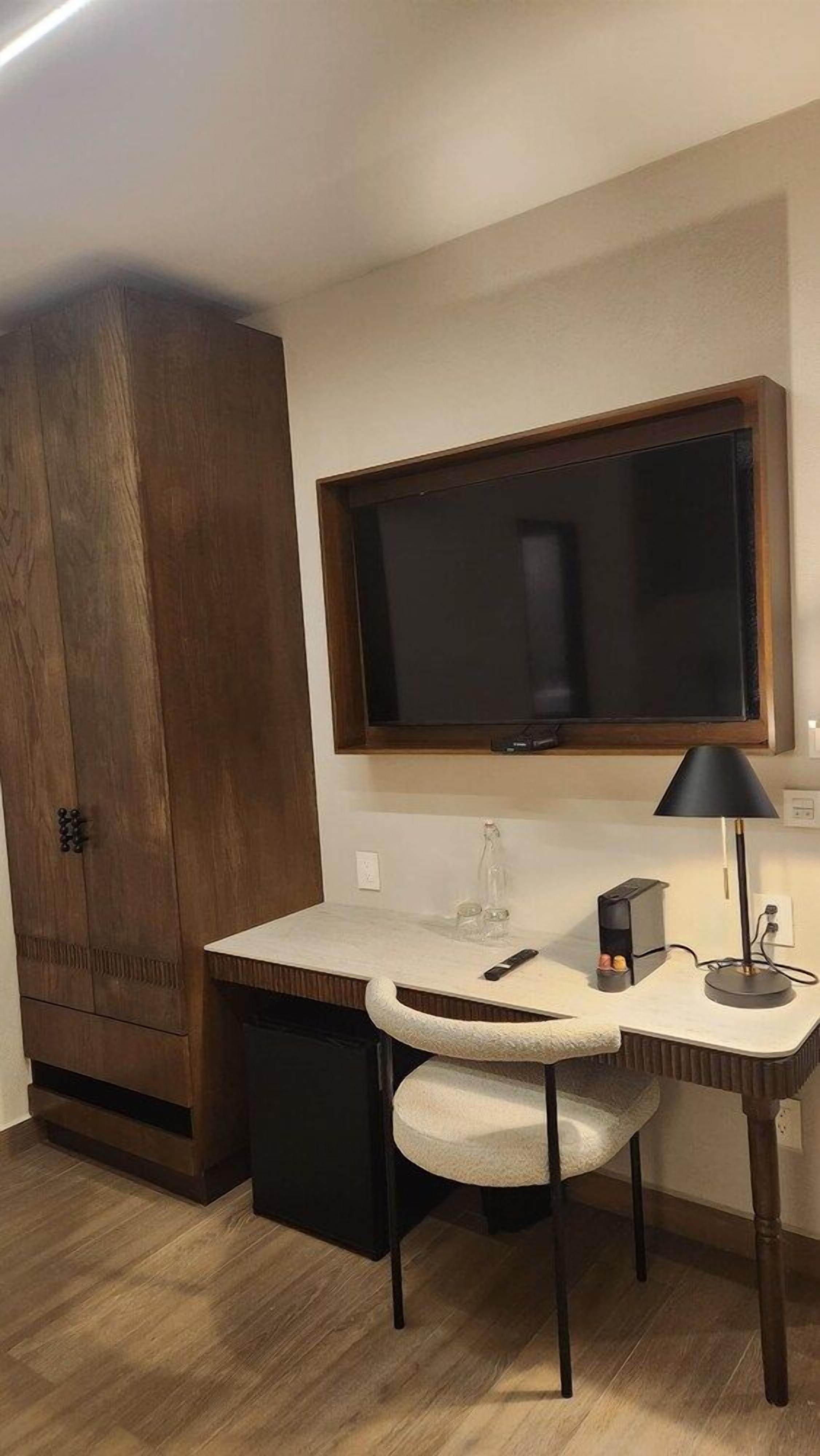 A compact room with a wooden wardrobe, a small desk with a chair, a television mounted above the desk, and a lamp on the desk.