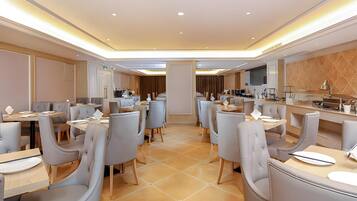 A modern hotel dining area with multiple tables and chairs, a kitchen in the background, and a tiled floor.