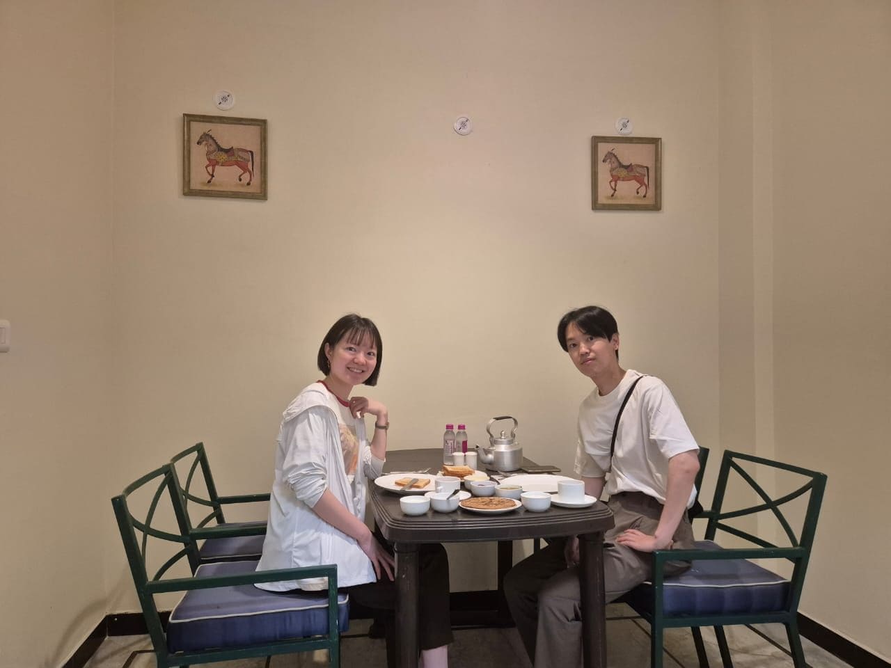 Two people seated at a table with plates, cups, and a teapot, in a room with chairs and framed pictures on the wall.
