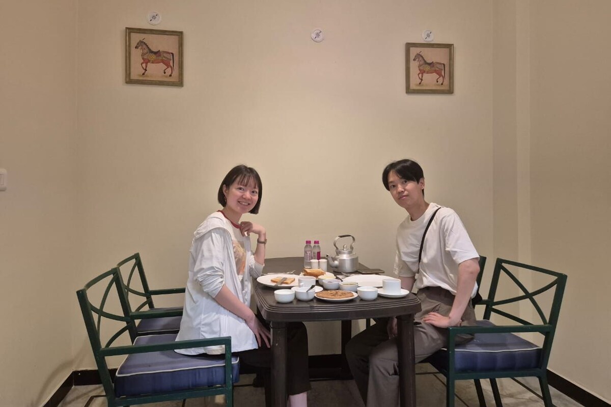 Dos personas sentadas a una mesa con platos, tazas y una tetera, en una habitación con sillas e imágenes enmarcadas en la pared.